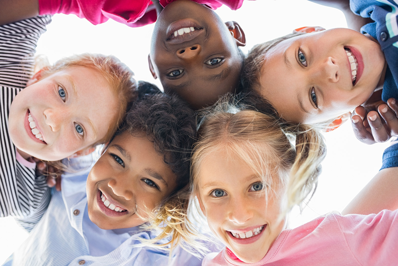 The image shows a group of smiling, happy children huddled together in a close circle, looking down at the camera. There are five children of various ethnicities, including both boys and girls, suggesting diversity and inclusivity. The children appear to be young, possibly in their elementary school years. They are wearing casual, colorful clothing and have bright, cheerful expressions on their faces. The image is shot from a low angle, giving the viewer the perspective of looking up at the children's faces as they peer down into the camera with joy and enthusiasm. The composition creates a sense of unity, friendship, and positive energy among the group of children.