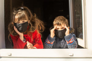Two children wearing masks look out a window.
